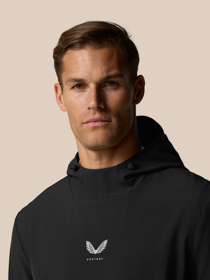 Close up of a man wearing a black Golf Pinnacle Performance Hoodie by Castore with a centered chest logo against a beige background.