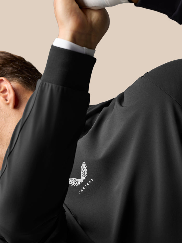 Close-up of a black golf performance hoodie with a white shirt cuff showing, featuring the Castore logo on the chest.