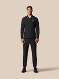 Full-body view of a man wearing a black Castore Golf Pinnacle LVS Winter Jacket with front zip against a beige backdrop