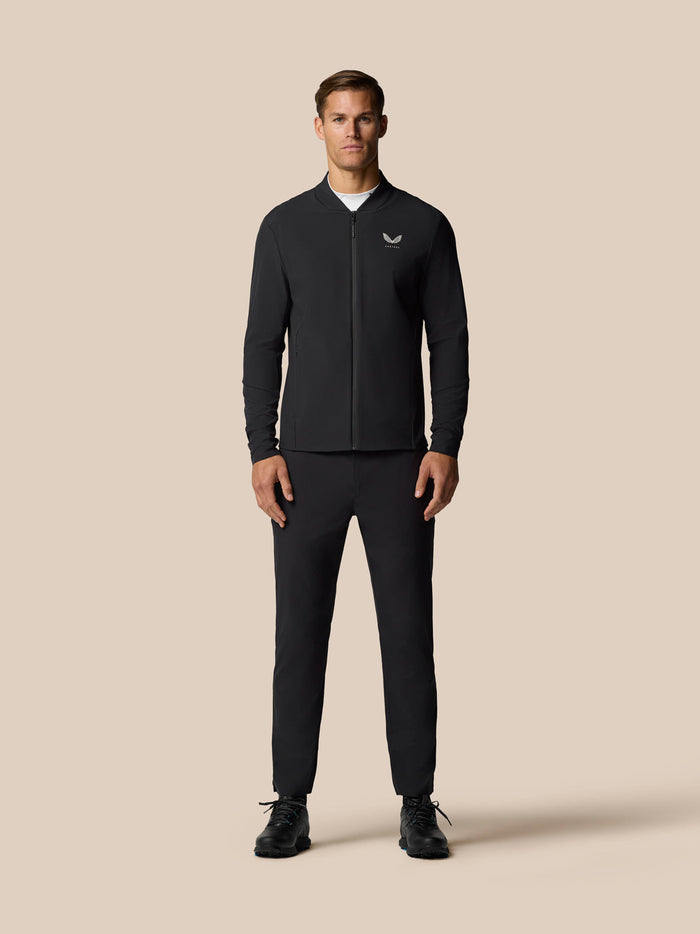 Full-body view of a man wearing a black Castore Golf Pinnacle LVS Winter Jacket with front zip against a beige backdrop