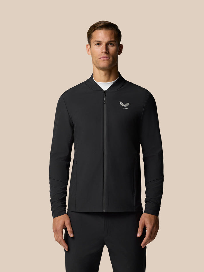 Full-body shot of a man in a black Castore Golf Pinnacle LVS Winter Jacket with front zip and left chest logo against a beige backdrop