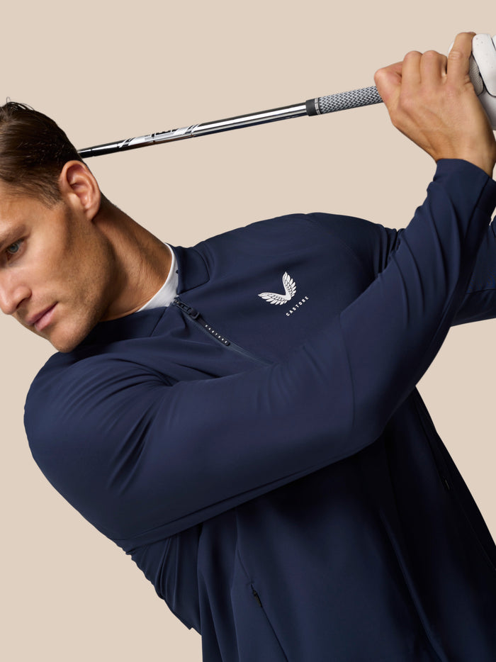 Male model wearing Midnight Navy Golf Pinnacle LVS Winter Jacket swinging a golf club in a studio against a beige background.