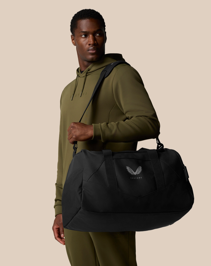 Male model in an olive hoodie carries a black Core Holdall duffel bag with logo.