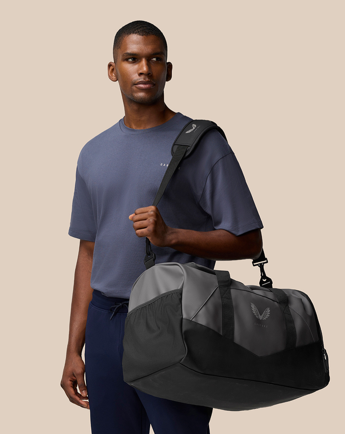 Male model in a blue gray tee holds a two tone gray and black Core Holdall with logo.