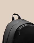 Close-up of a black CORE BACKPACK with a sturdy top handle and curved zipper against a beige background