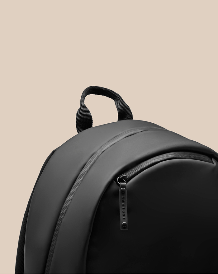 Close-up of a black CORE BACKPACK with a sturdy top handle and curved zipper against a beige background